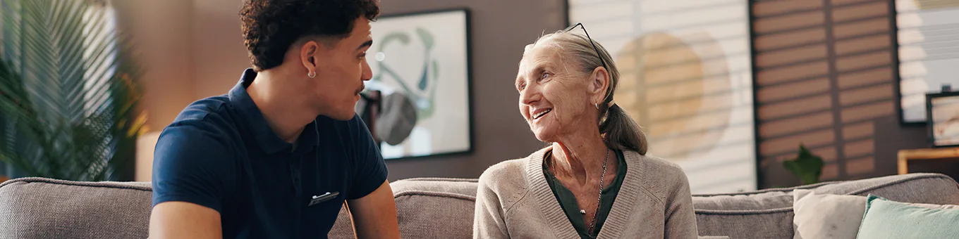 Betreuungskraft sitzt mit Patienten auf dem Sofa und unterhalten sich.